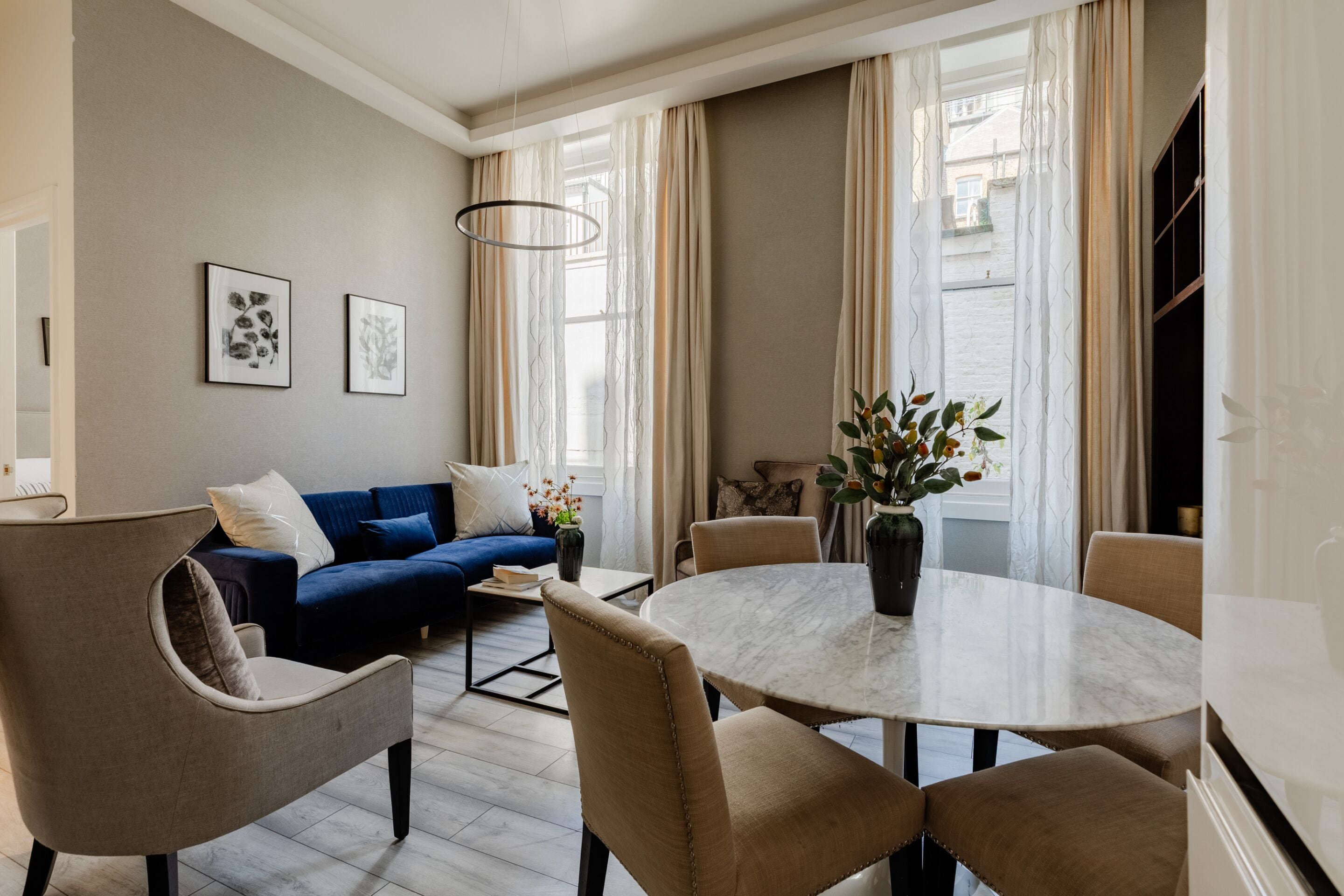 Et moderne spiseområde med et rundt bord, beige stole, en blå sofa og en vase med blomster.