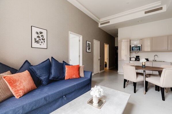A modern living room with a blue sofa, orange and blue cushions, a white coffee table with a vase, and a dining area with a wooden table and chairs.
