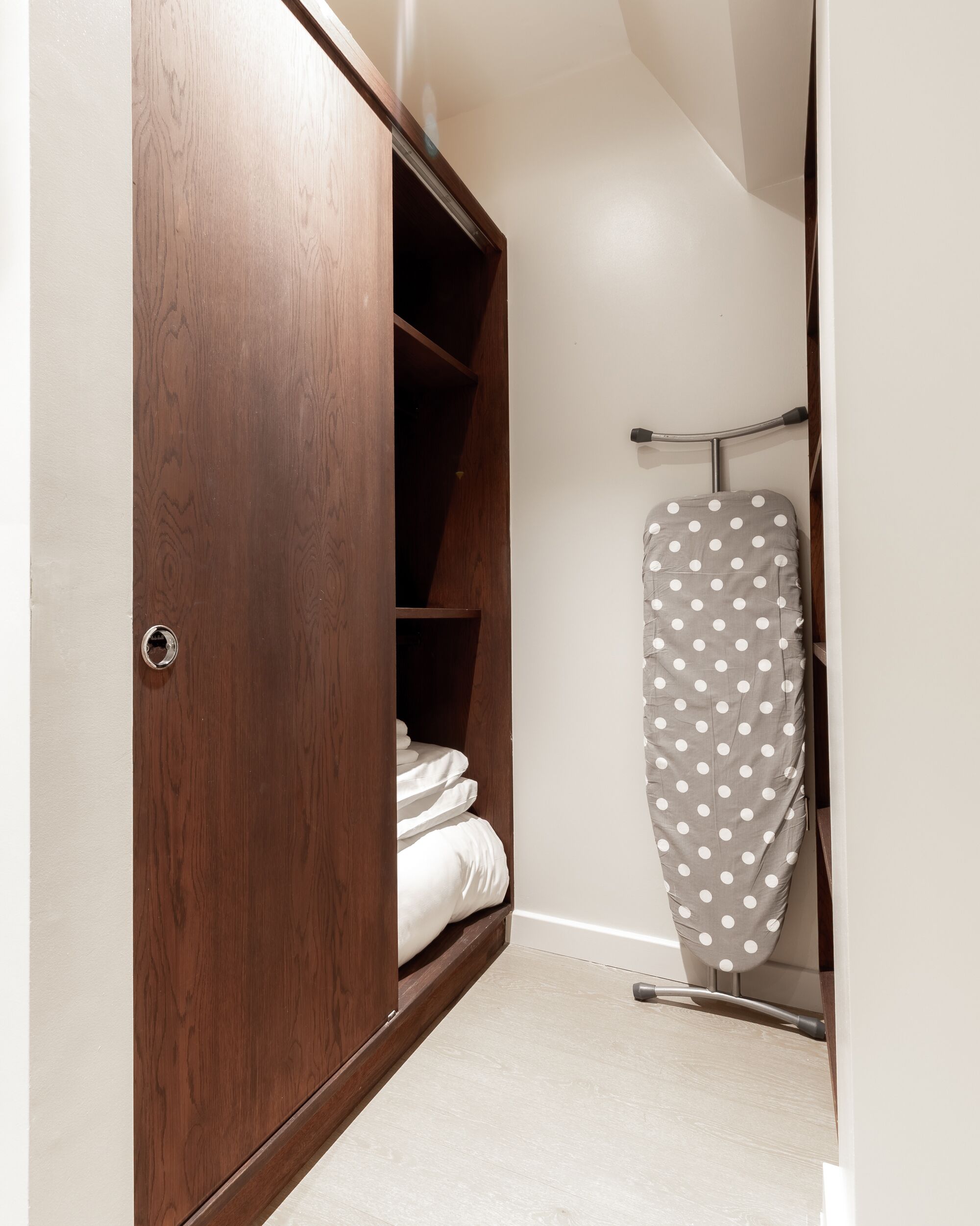 A closet with wooden doors, a metal ironing board, and neatly folded white linens.