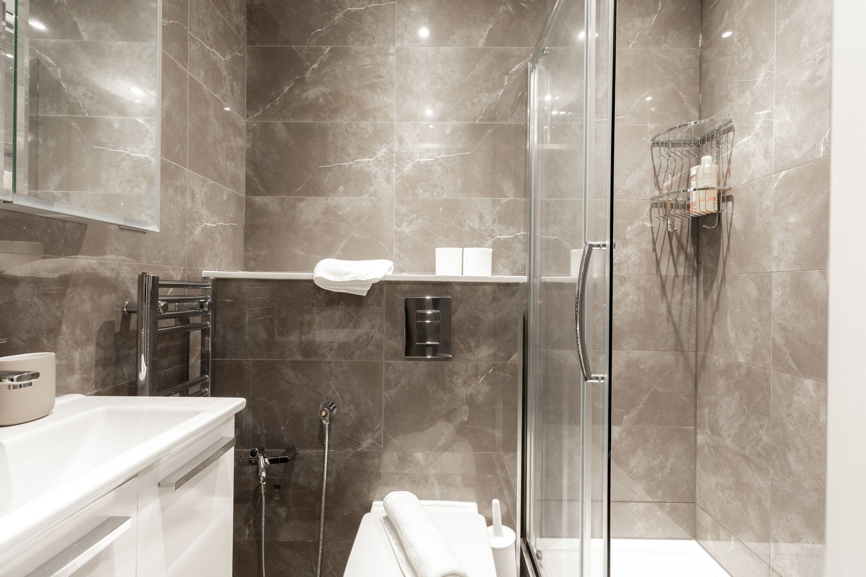 A modern bathroom with a glass shower enclosure, marble tiles, a white sink, and a towel rack.