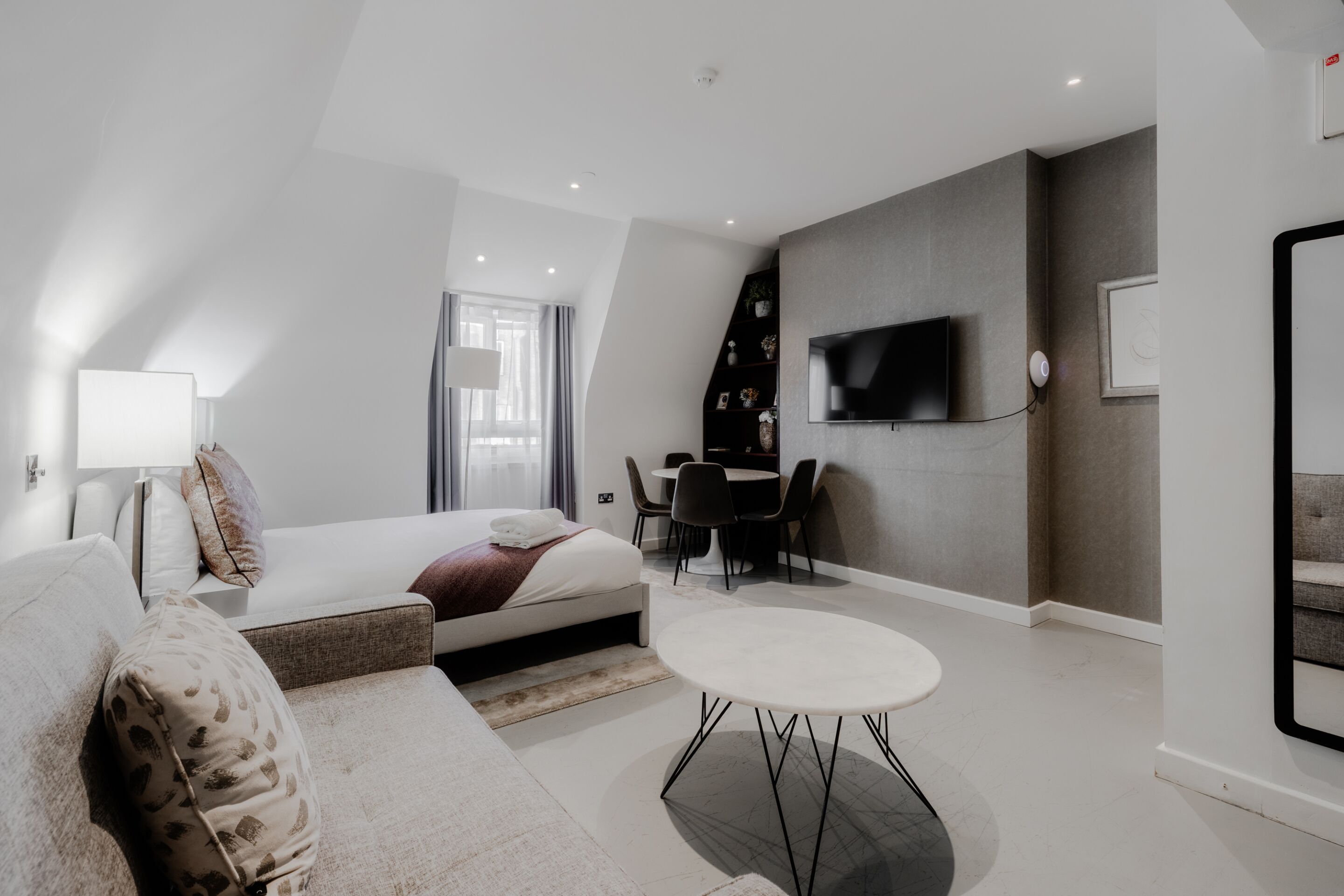 A modern hotel room with a bed, a sofa, a dining table, and a TV mounted on the wall.