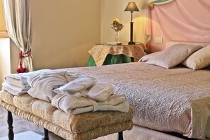 A neatly made bed with a patterned bedspread, a stack of folded towels on a bench, a bedside table with a lamp and a vase of flowers. - Signoria Farine (Florence)