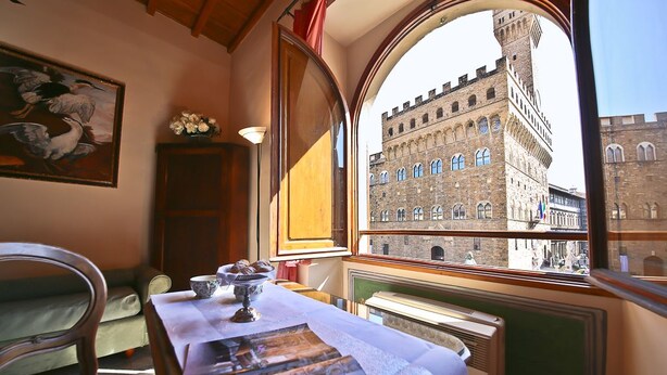 A room with a table covered in newspapers, a painting of birds, and a view of a historic building through a window. - Signoria Farine (Florence)