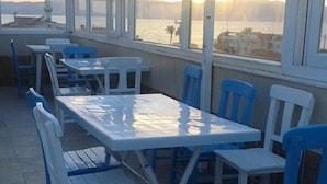 Ett utomhusområde med sittplatser, vita bord och blå stolar, med utsikt över en vattenyta och ett avlägset landskap.