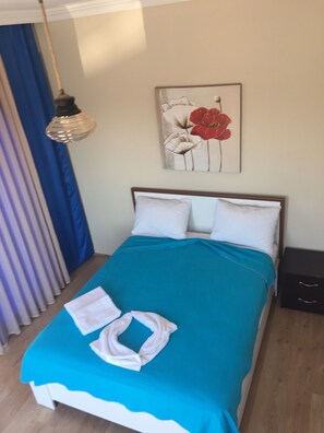 Una cama bien hecha con una colcha azul y almohadas blancas, un cuadro de flores rojas en la pared, una cortina azul y un suelo de madera.