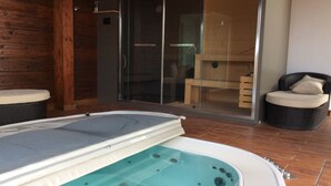 Un área de spa con hidromasaje, suelo de madera, ducha envuelta en cristal y una silla de mimbre.