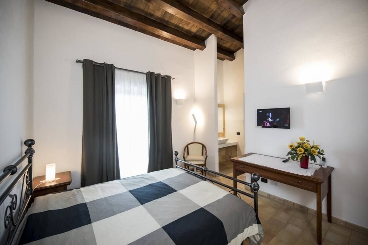 A bedroom with a bed, a nightstand, a chair, a table with a vase of flowers, and a TV on the wall.