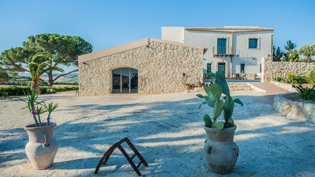 Un edificio de piedra con una amplia entrada arqueada, un patio con plantas en macetas y una vista despejada de un edificio más grande al fondo.