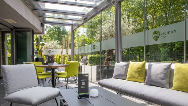Un espace extérieur moderne, aménagé pour se détendre, avec une structure en verre, un canapé avec des coussins et une table portant un panneau avec un code QR.