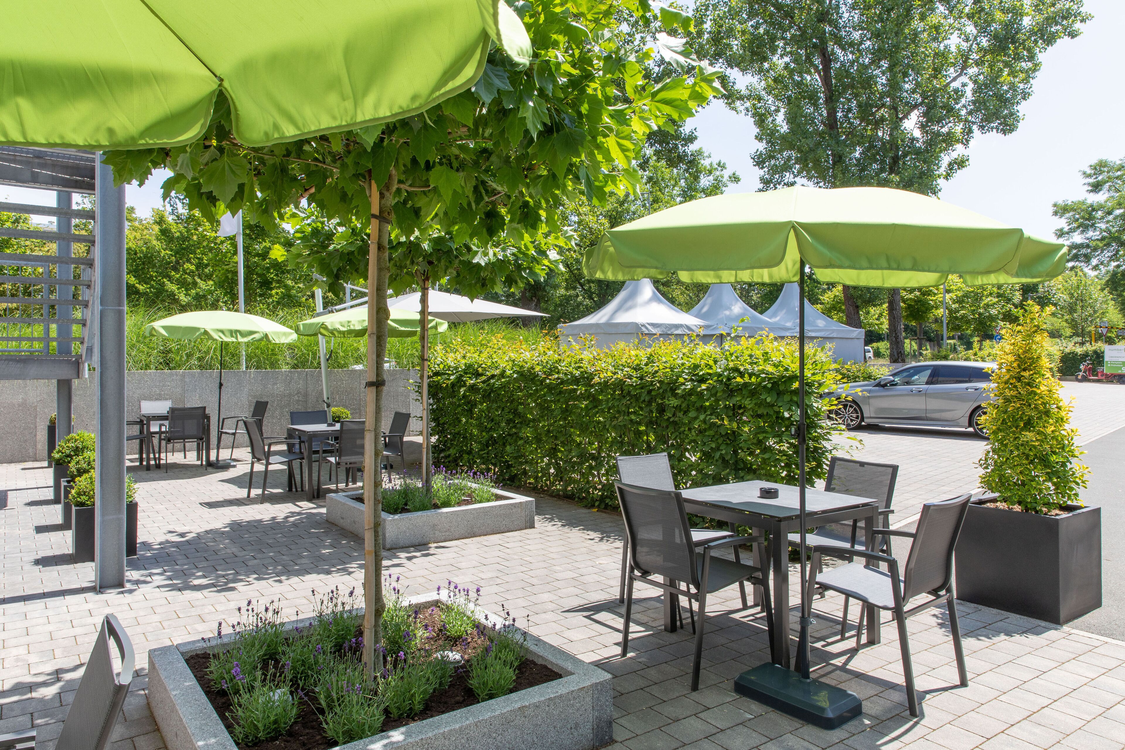 An outdoor patio area with tables and chairs under green umbrellas, surrounded by planters and a hedge.