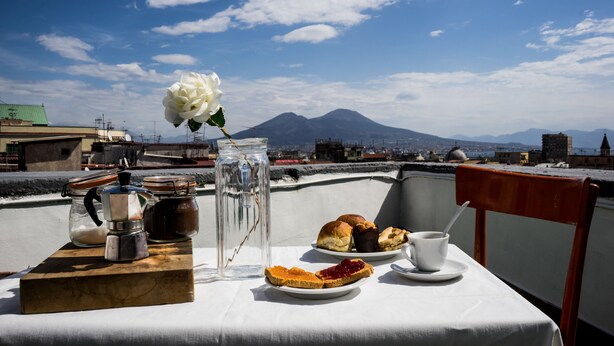 A table with food and drink items on a balcony overlooking a cityscape with a mountain in the background. - Taggarb B&B (Naples)