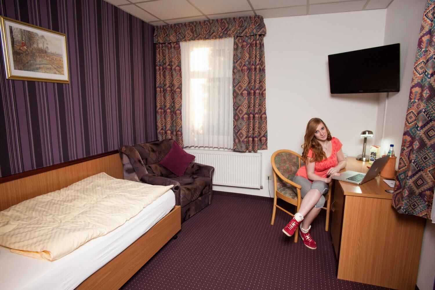 A hotel room with a bed, a sofa, a desk with a laptop, a TV, and patterned curtains.