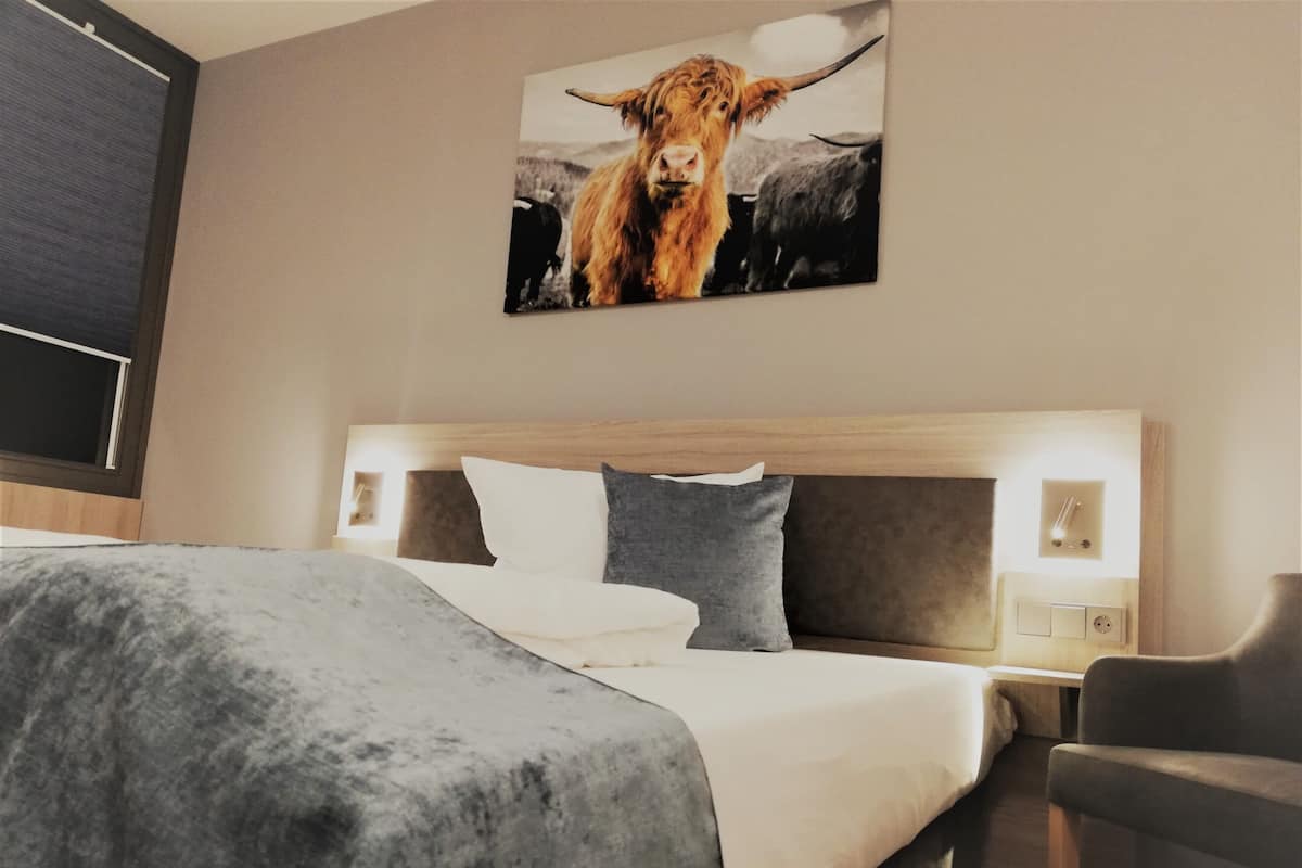 A hotel room with a large bed, a headboard with integrated lighting, a grey blanket, and a painting of a cow on the wall.