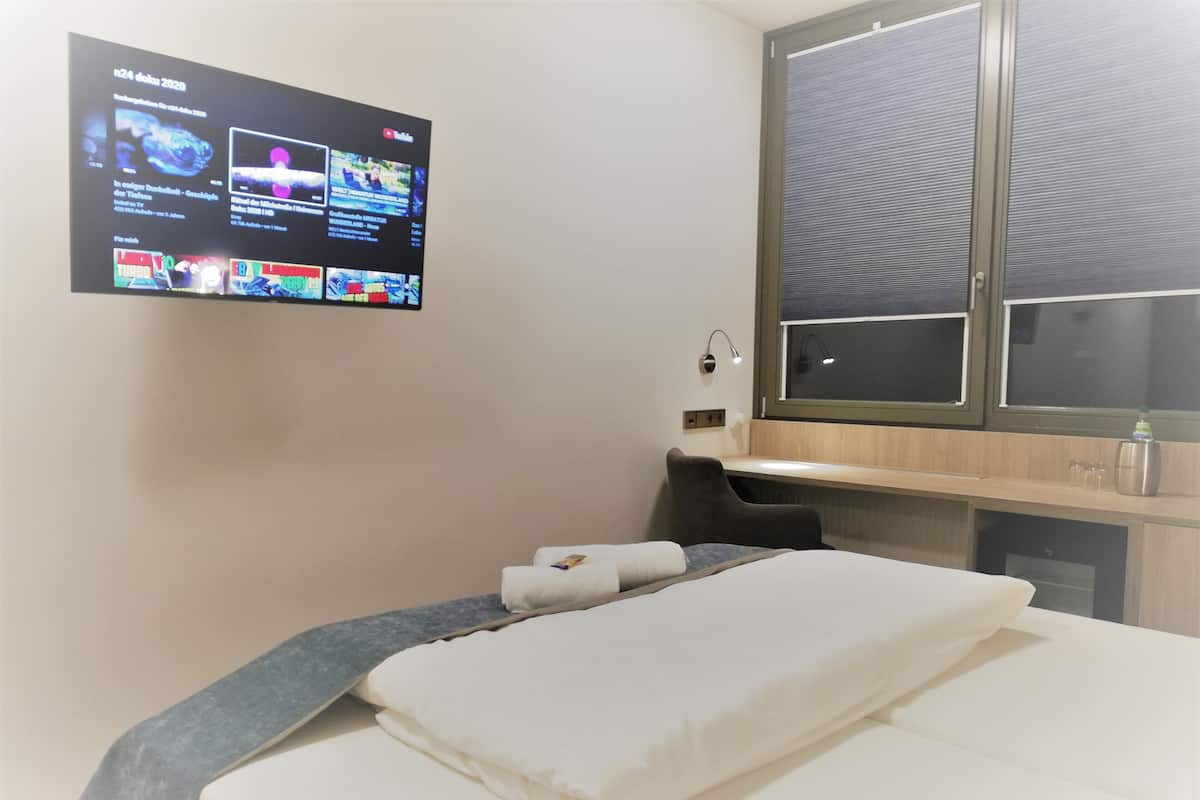 A hotel room with a bed, a desk, a TV mounted on the wall displaying various channels, and a window with blinds.
