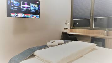 Habitación de hotel con cama, escritorio, televisor montado en la pared mostrando varios canales y ventana con persianas.