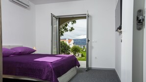 A bedroom with a bed, purple bedding, a view through an open door to a balcony, and a white wall. - Apartments Bago (Split)
