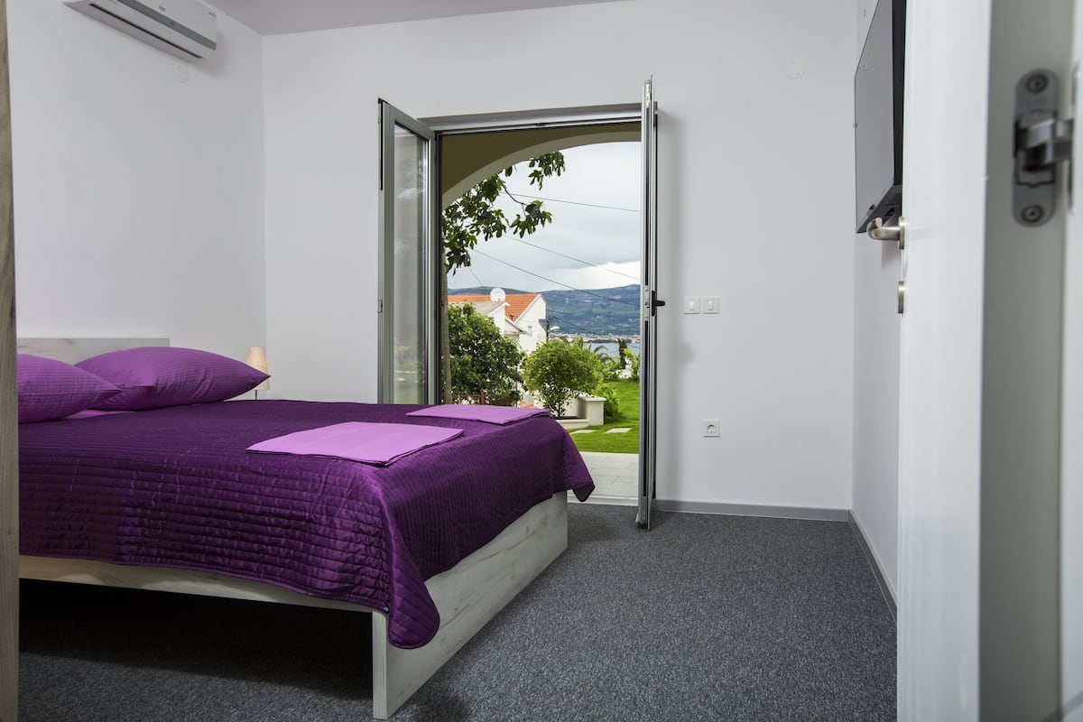 A bedroom with a bed, purple bedding, a view through an open door to a balcony, and a white wall.