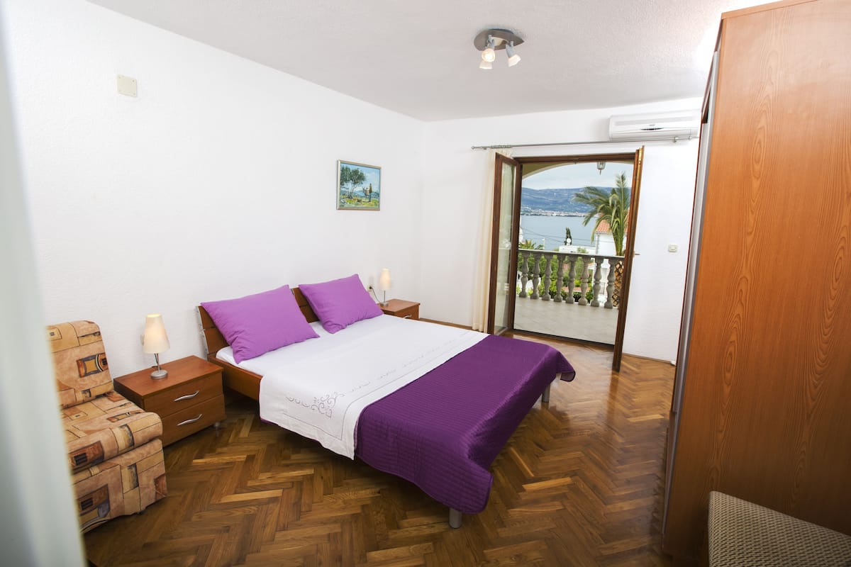 A bedroom with a bed, nightstands, a wooden floor, a balcony with a view, and a painting on the wall.