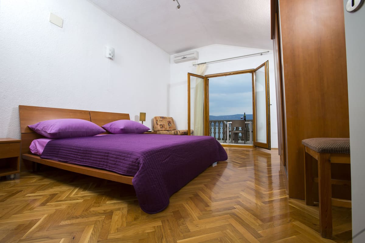 A bedroom with a wooden bed, a view of the city through a large window, and a balcony with a chair.