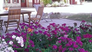 Une cour avec une table et des chaises, entourée de fleurs aux couleurs vives et d’un bâtiment dont les fenêtres et les portes sont encadrées de rouge.