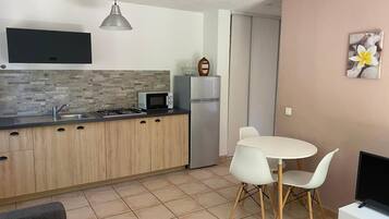 Een kleine keuken met een magnetron, fornuis en koelkast, een eethoek met een witte tafel en stoelen, en een televisie op een witte kast.