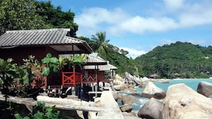 A traditional wooden building with a tiled roof, located near a rocky shoreline with a view of the ocean and a lush green hill in the background. - OK2 Bungalow (Koh Tao)