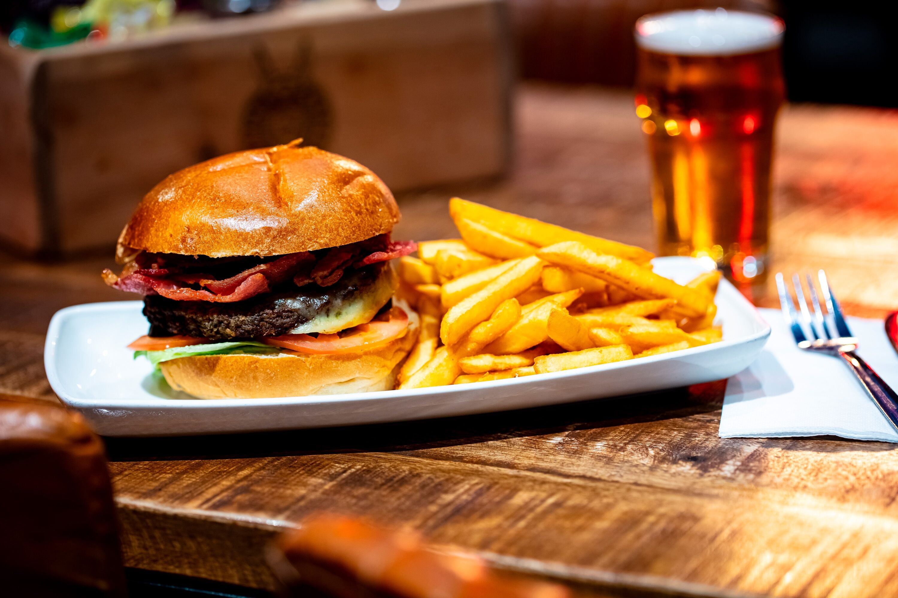 A plate with a burger and fries, a glass of beer, and a fork on a wooden table.