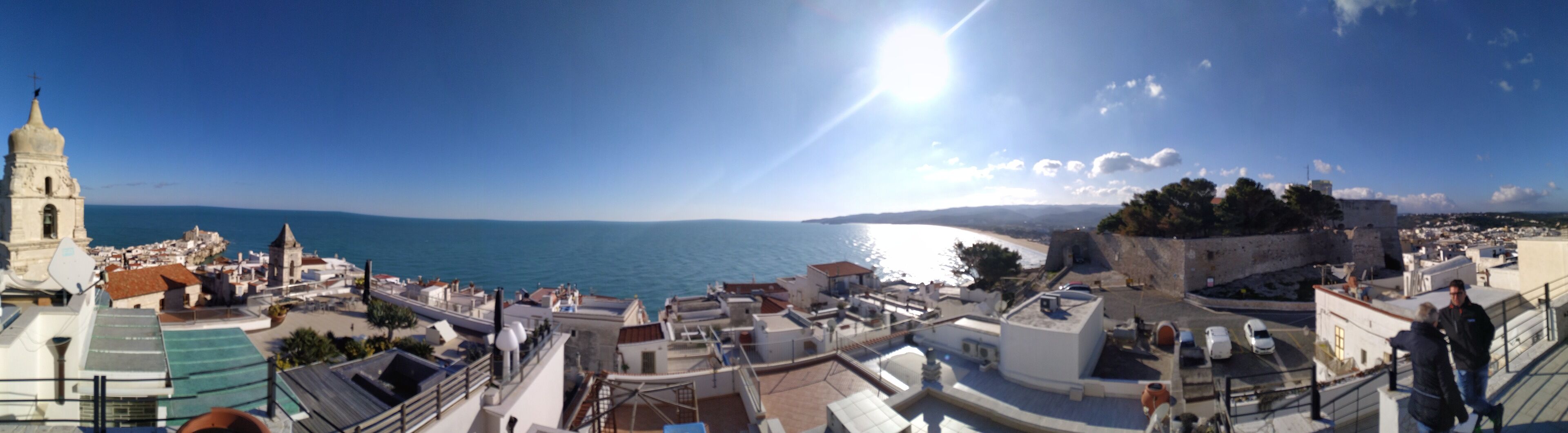 Vista panorâmica de uma cidade costeira com uma igreja imponente, uma muralha fortificada e uma visão clara do mar.