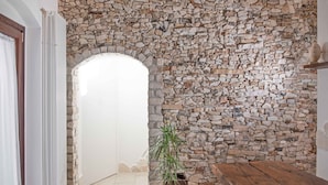 Uma parede de pedra com um arco que dá acesso a um aposento, uma planta em vaso e um banco de madeira.