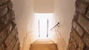 Uma escadaria de pedra leva a um quarto com teto branco e janela no alto.
