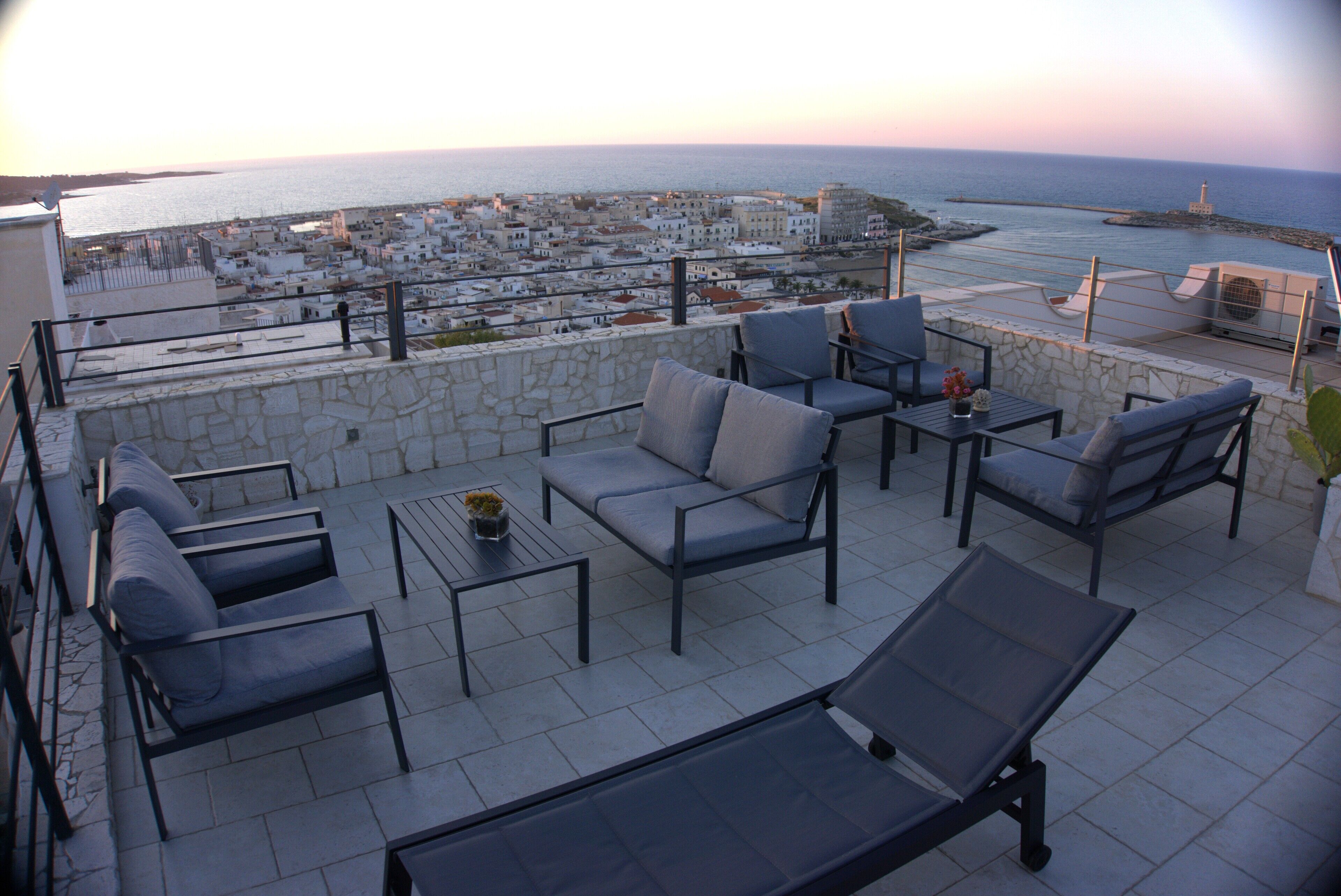 A terrace with outdoor seating, a stone wall, and a view of the sea and buildings.