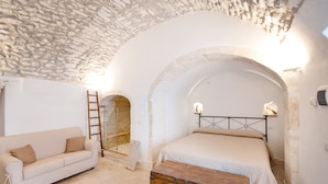Quarto com teto de pedra, um banco de madeira, cama com cabeceira de metal e sofá branco.