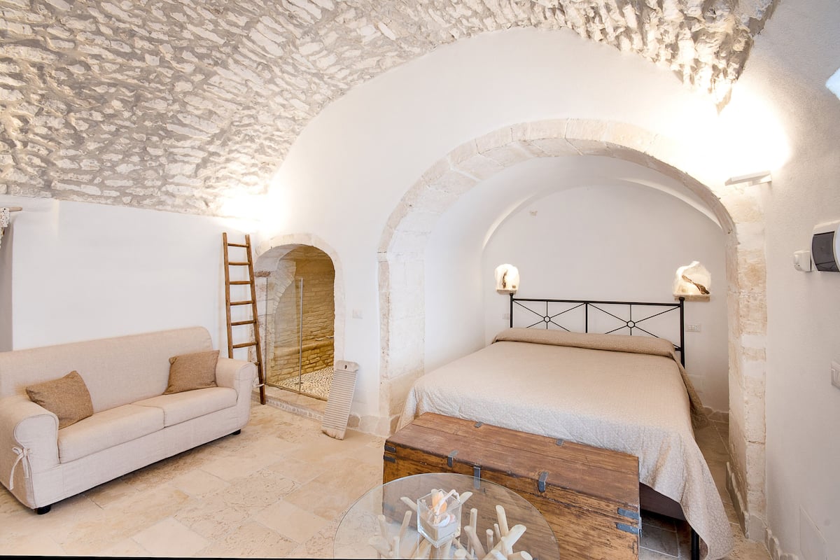 Ein Schlafzimmer mit Steindecke, einer Holzbank, einem Bett mit Metallkopfteil und einem weißen Sofa.