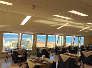 A dining area with tables and chairs, large windows with a view of the beach and ocean, and ceiling lights. - Hotel Borgo Marina (Rodi Garganico)