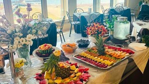 Une table garnie d’une variété de fruits et d’un gâteau en forme de pastèque, le tout mis en valeur par une vue sur la plage en arrière-plan.
