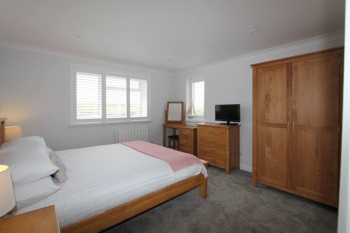 Een slaapkamer met een bed, dressoir, televisie, kledingkast en ramen.