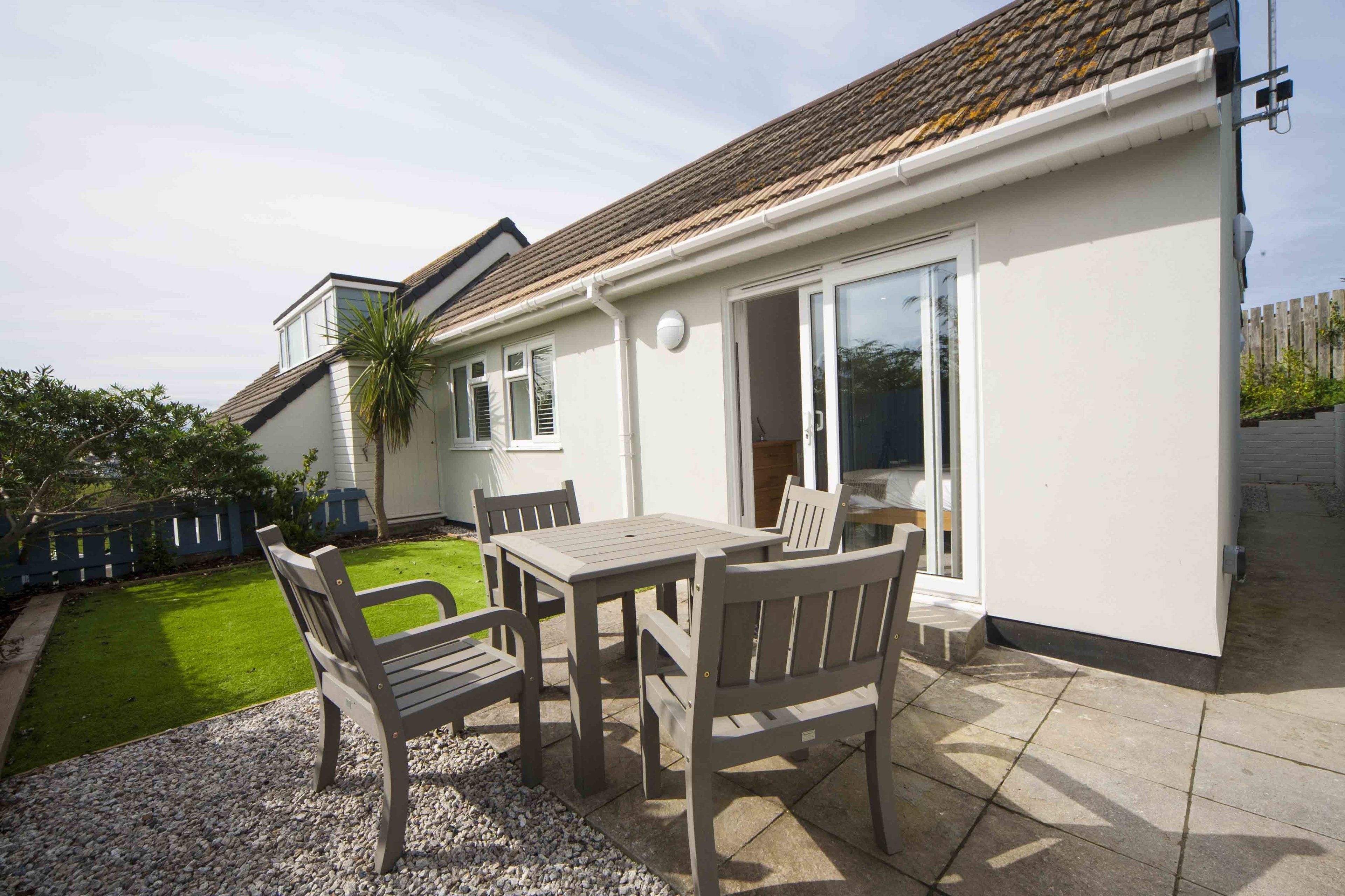 A patio with a table and chairs, a small garden, and a house with a tiled roof and white exterior.