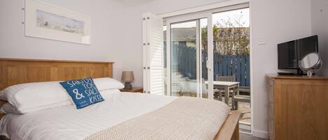 Une chambre à coucher comprenant un lit, une tête de lit en bois, une table de chevet avec une lampe, un téléviseur posé sur un meuble en bois, et une porte-fenêtre coulissante donnant sur un espace extérieur.