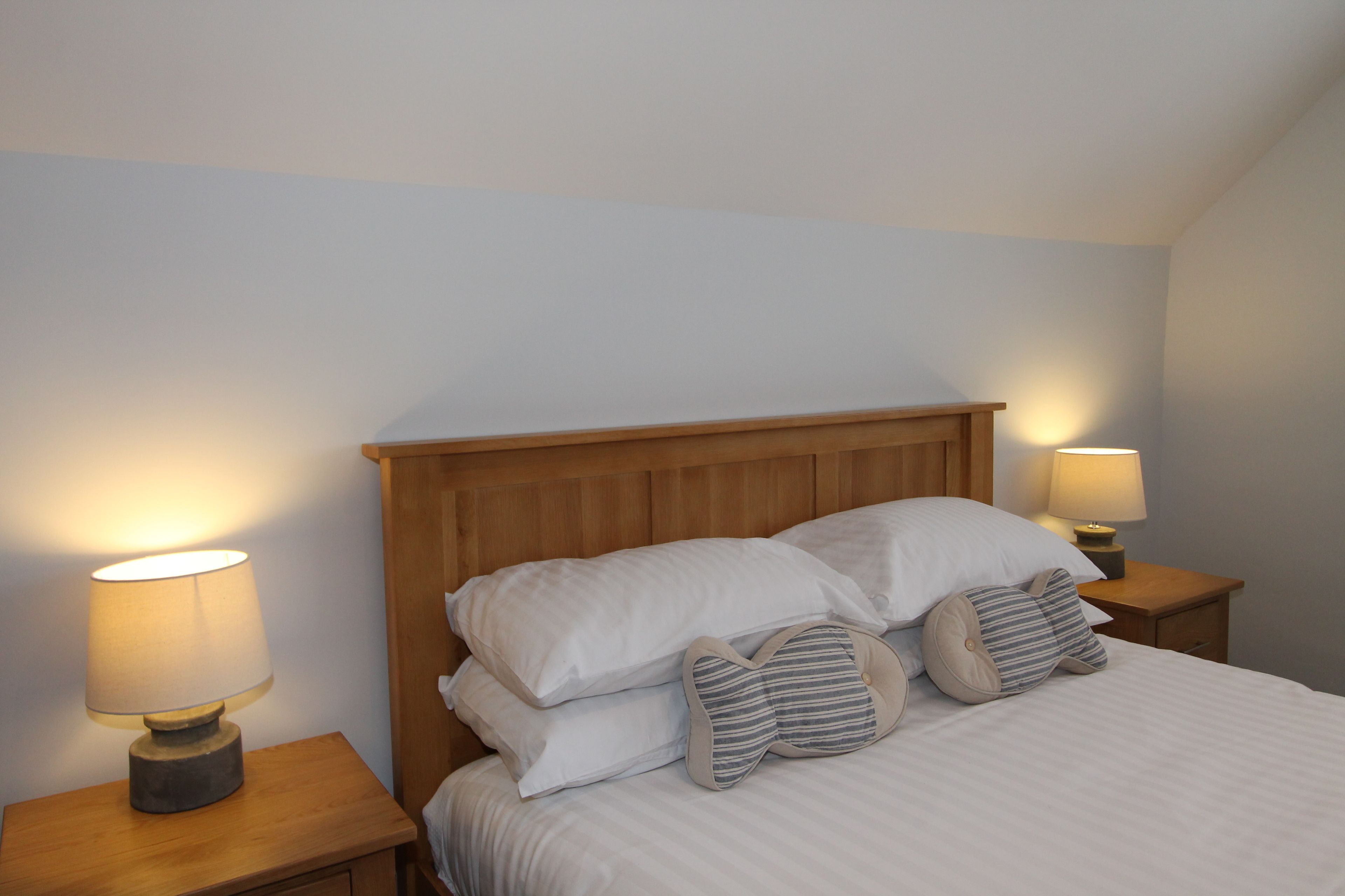 Cama bem arrumada com lençóis brancos e almofadas listradas, com duas luminárias de cabeceira sobre mesas de cabeceira de madeira.