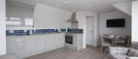 Een moderne keuken met witte kasten, een blauwe achterwand en een ingebouwde oven. Er is een eethoek met een tafel en stoelen, en een zithoek met een bank en salontafel.