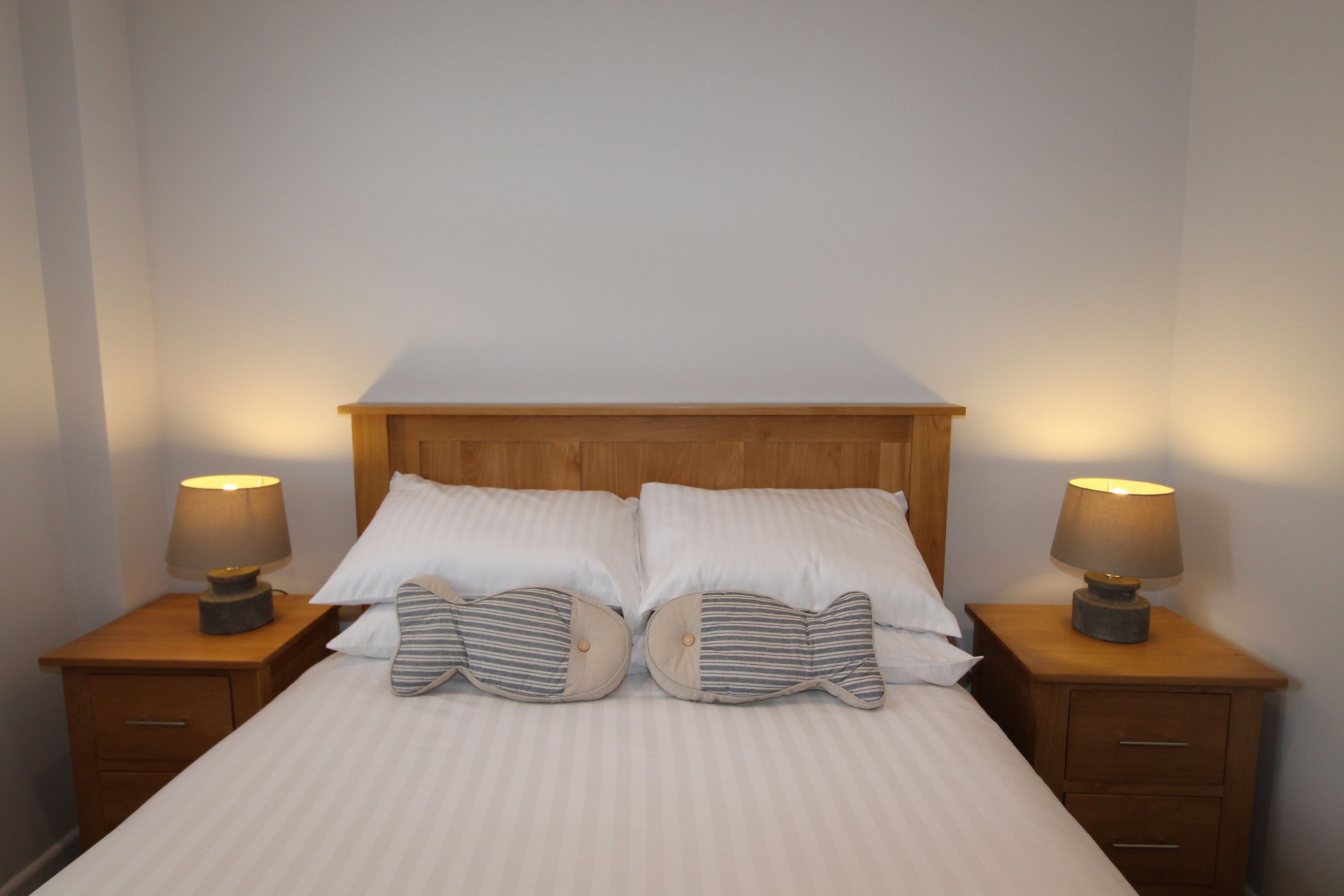 Una cama bien hecha con sábanas blancas y almohadas a rayas, con dos mesitas de noche con lámparas, en una habitación sencilla con cabecera de madera y paredes lisas.