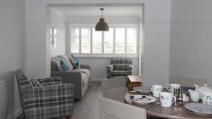 Een moderne woonkamer met een bank, twee fauteuils, een eettafel met theeservies en uitzicht door grote ramen.