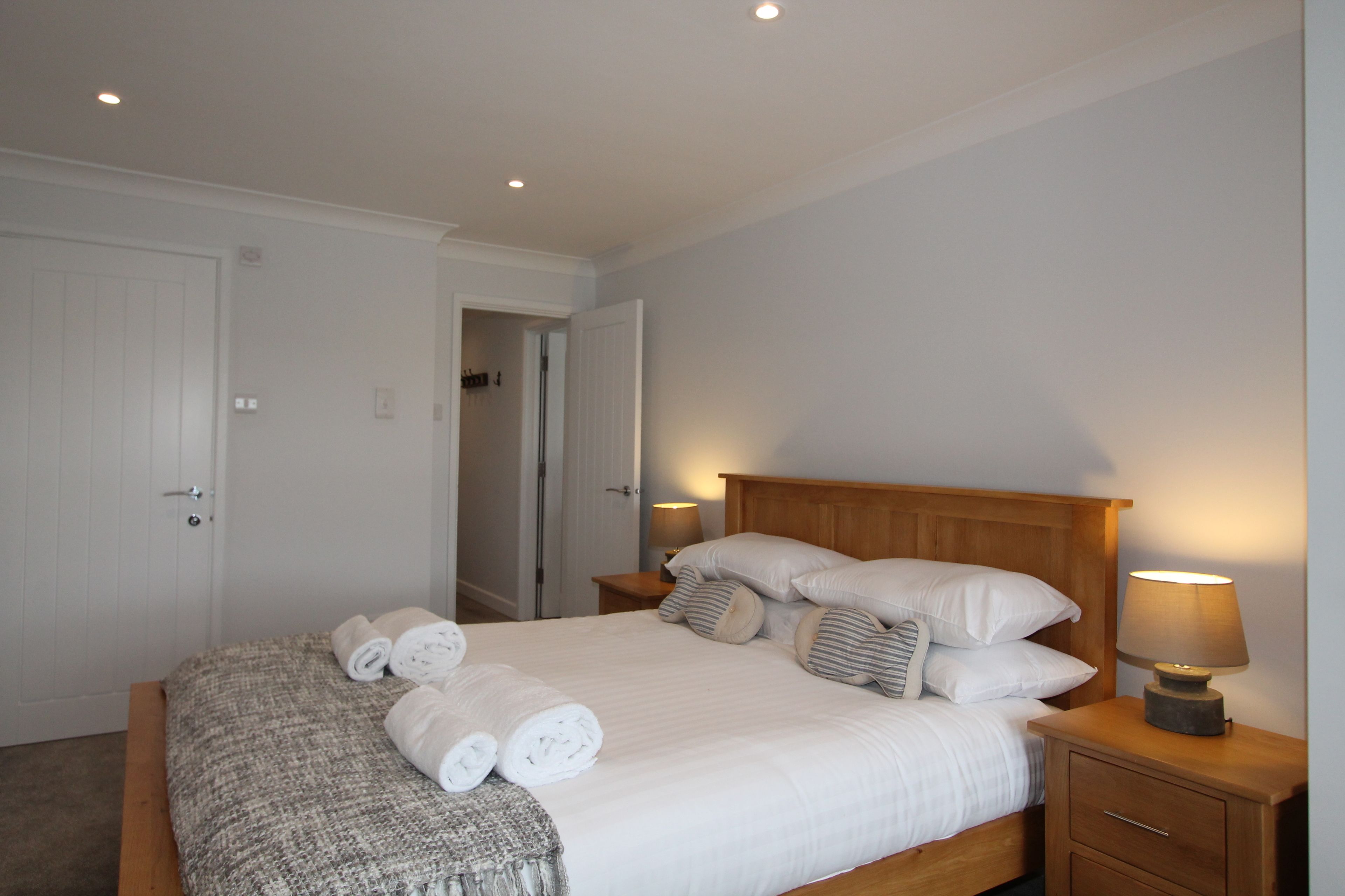 Una cama bien tendida con almohadas y una lámpara de noche, una mesita de noche de madera y una puerta a otra habitación.