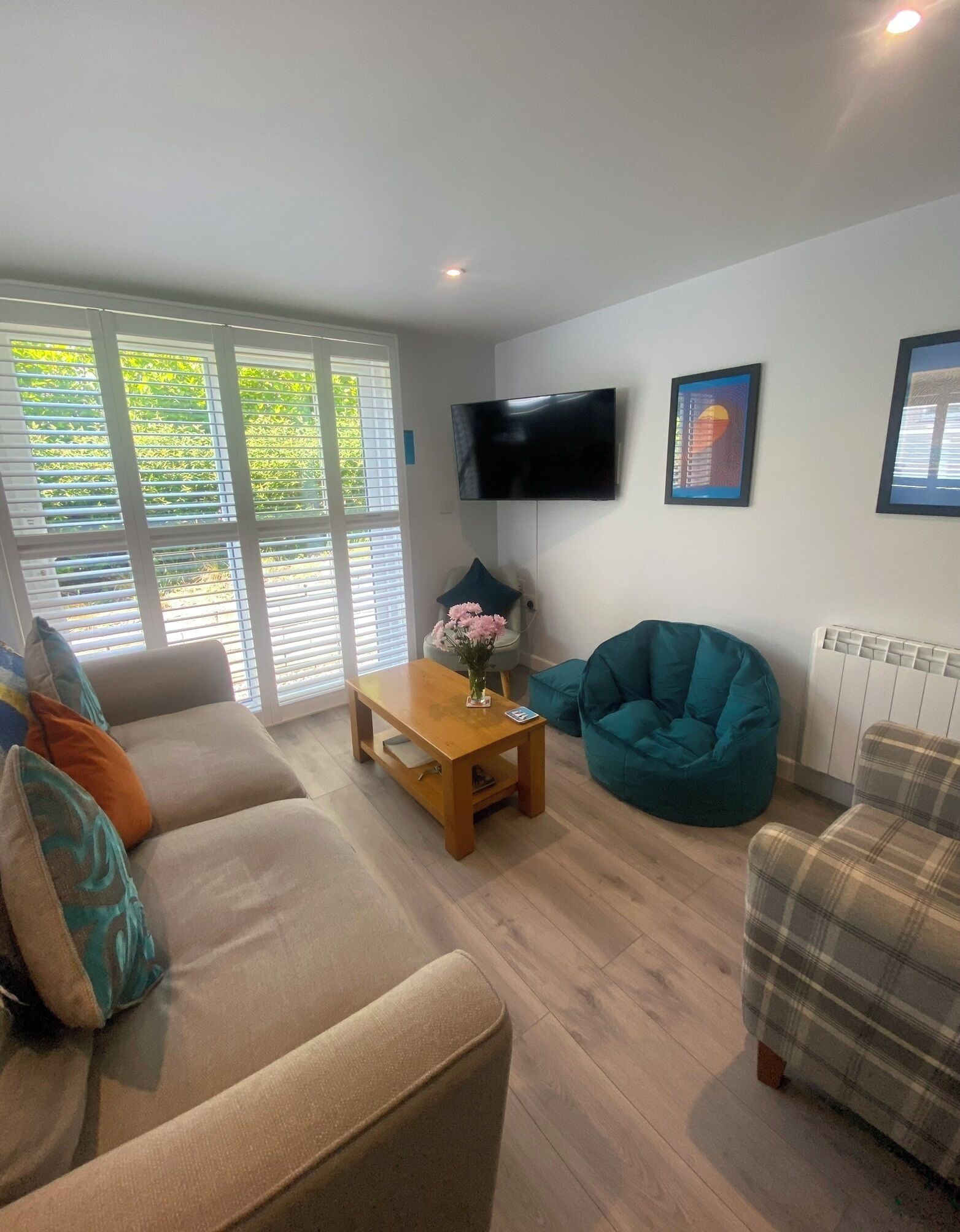 Un salon avec un canapé, une table basse, un pouf vert turquoise et un fauteuil à carreaux. Un téléviseur à écran plat est fixé au mur.
