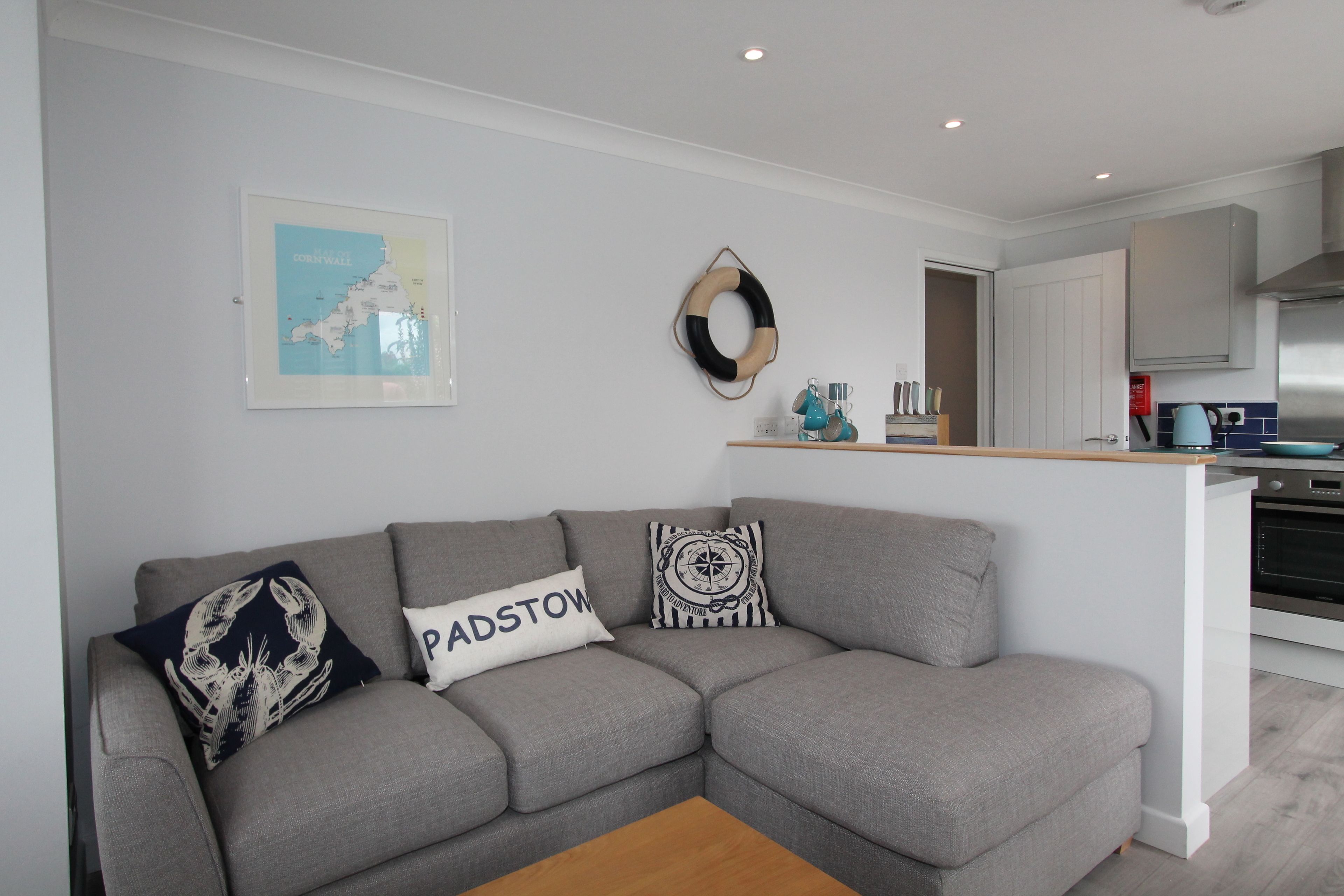 A modern living room with a grey sectional sofa, a wooden coffee table, a kitchen area with white cabinets, and a map on the wall.