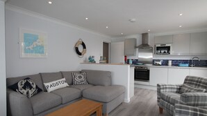 Een moderne woonkamer met een grijze bank, een houten salontafel en een geruit fauteuil. In de achtergrond is een keuken zichtbaar met witte kasten en een blauwe spatwand.