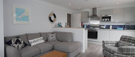 A modern living room with a grey sofa, a wooden coffee table, and a plaid armchair. A kitchen with white cabinets and a blue backsplash is visible in the background.