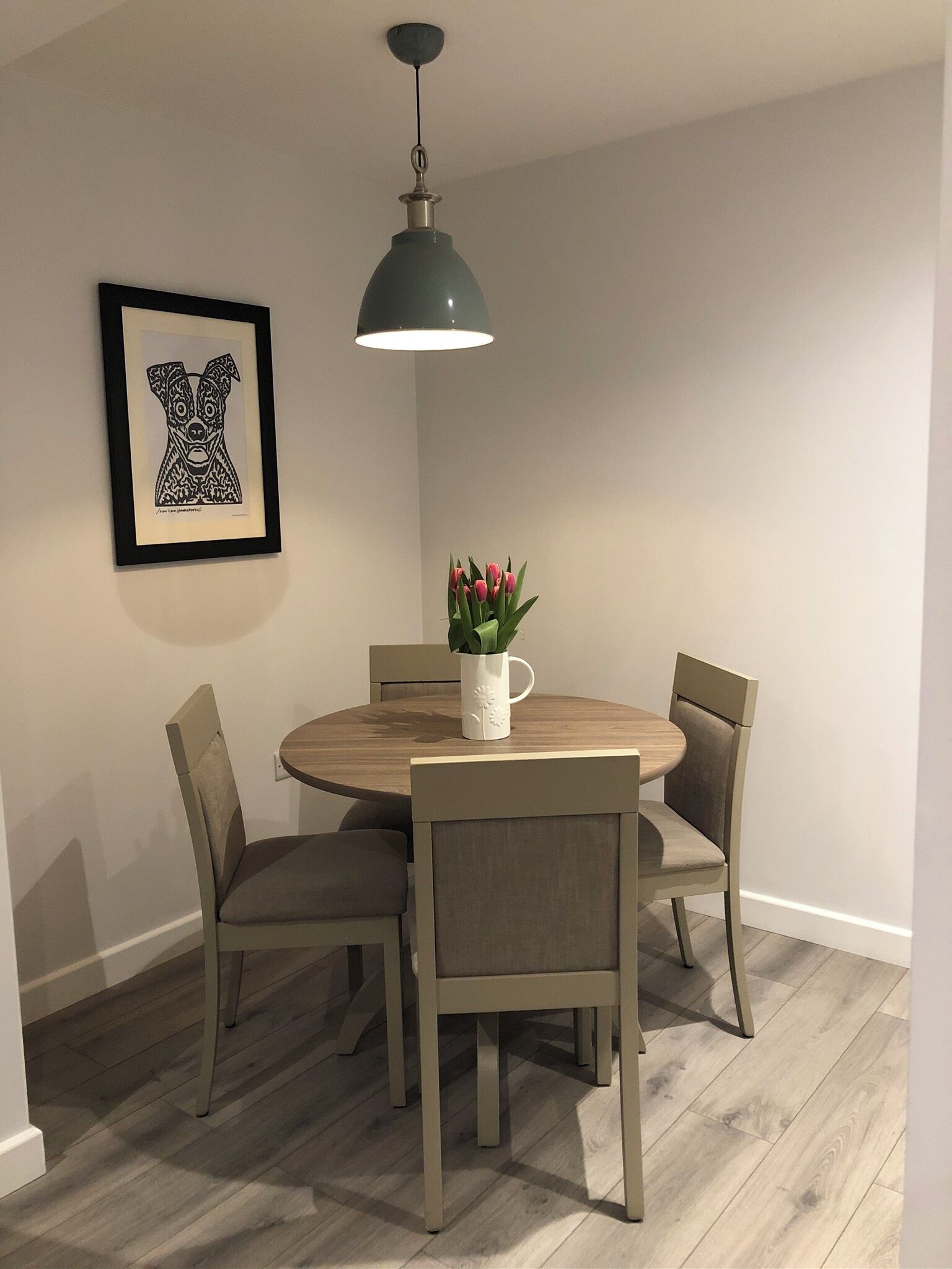 A dining area with a round table, four chairs, a pendant light, a framed artwork, and a vase with flowers.