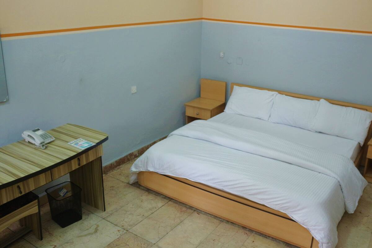 A bedroom with a single bed, a bedside table, a nightstand, a desk with a telephone, and a trash bin.