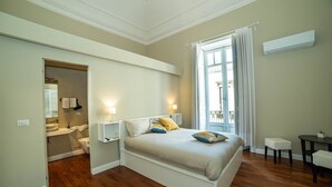 A bedroom with a bed, bedside tables, a window with curtains, a small table, and a bathroom visible through an open door. - Le 4 Leggende (Catania)